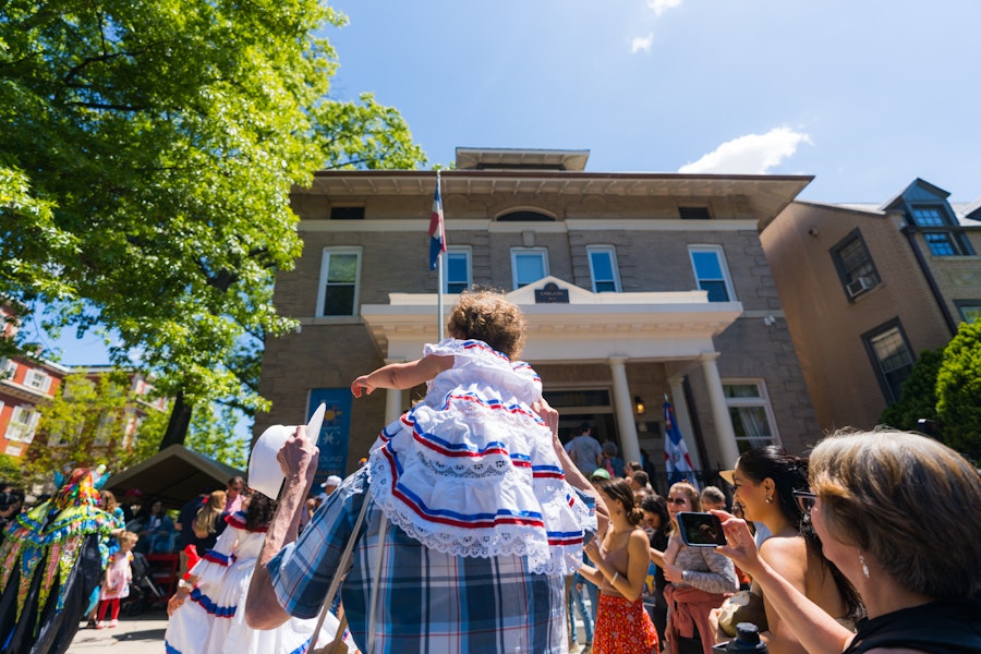 A little girl in a dress sits on the shoulders of a family member, watching performers outside of an embassy. 