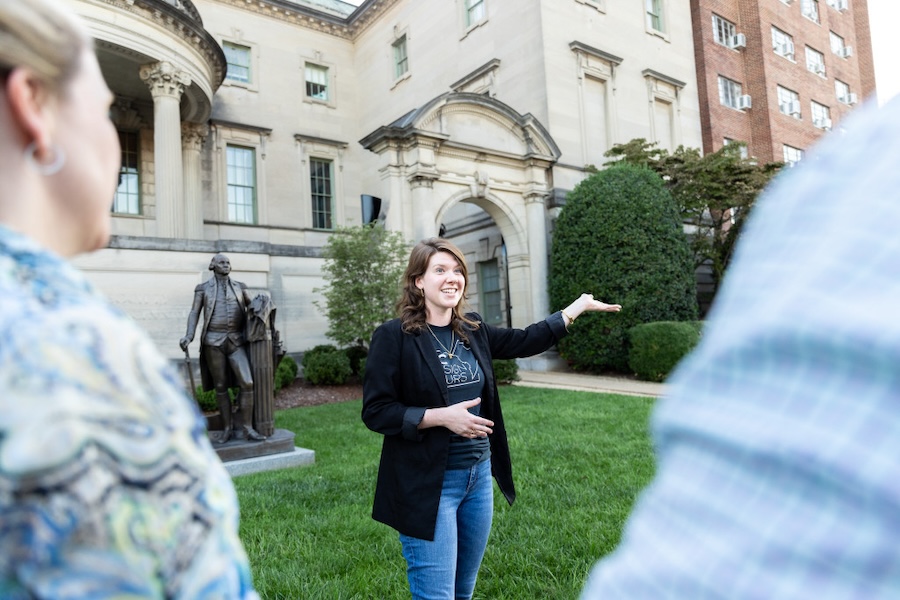 A tour guide gestures towards a historic building with a statue out front. 