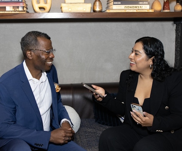 This image shows two people sitting on a couch, engaged in a friendly and professional conversation. The woman on the right is holding a phone, possibly conducting an interview. Both are smiling, suggesting a positive and engaging discussion. The setting appears casual yet professional, possibly a media or networking event.