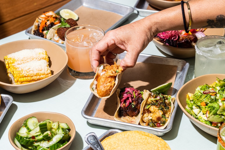A table filled with colorful tacos and small plates, with a hand reaching to grab a bite. 