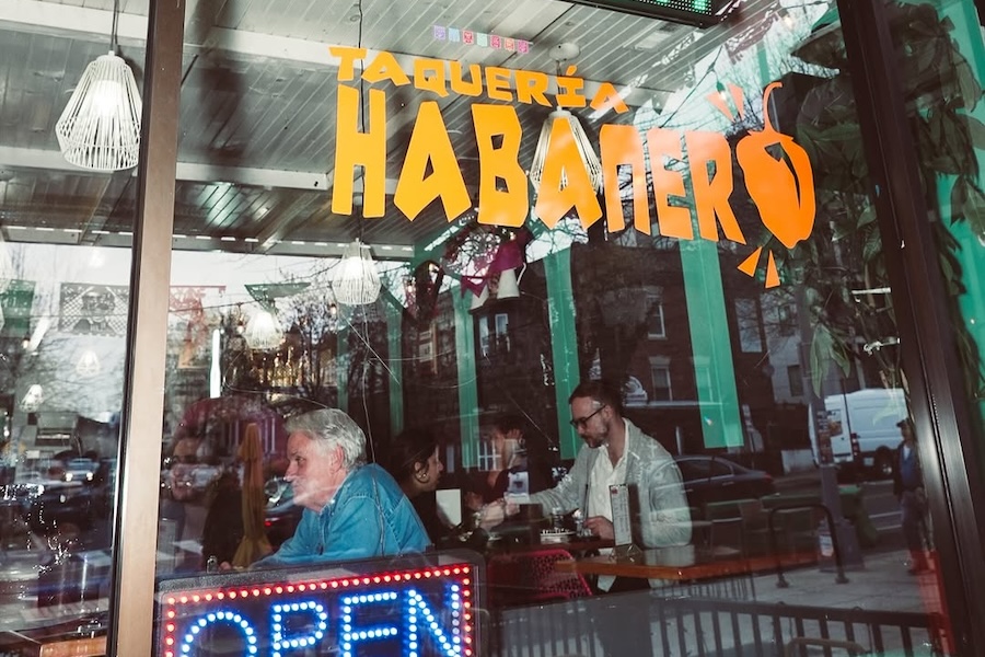 The exterior window of a restaurant (with people inside) that reads "Taqueria Habanero" in bright yellow and orange lettering. 