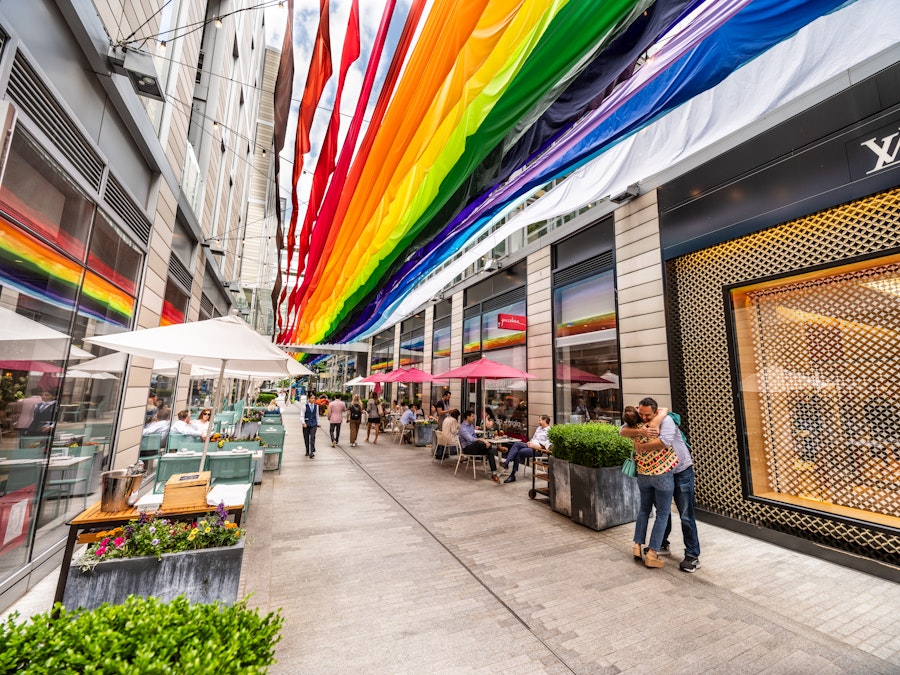 Colorful rainbow fabric banners hang above a lively pedestrian alley at CityCenterDC, where people dine outdoors and stroll between modern shops and restaurants.