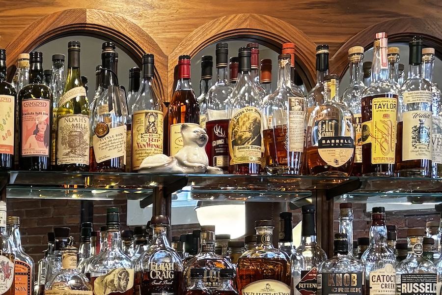 A mirrored bar shelf displays an array of whiskey bottles and a ceramic white cat figurine.