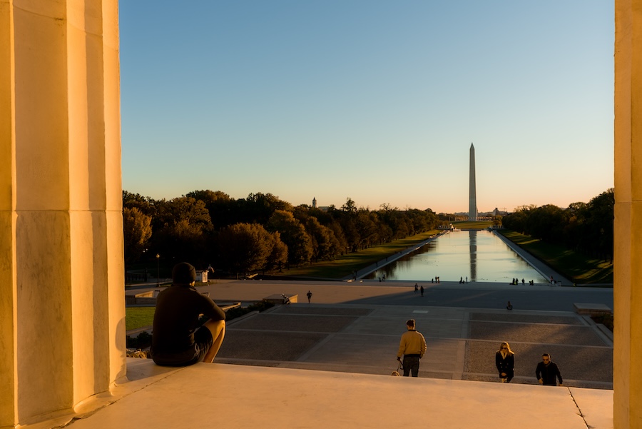 A person sits on the steps of the Lincoln Memorial overlooking the Reflecting Pool and Washington Monument at sunset.