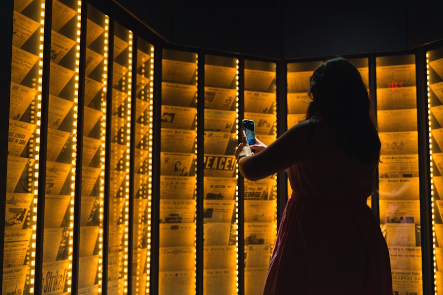 A visitor photographs a glowing wall of historic newspaper headlines in the Smithsonian National Postal Museum.