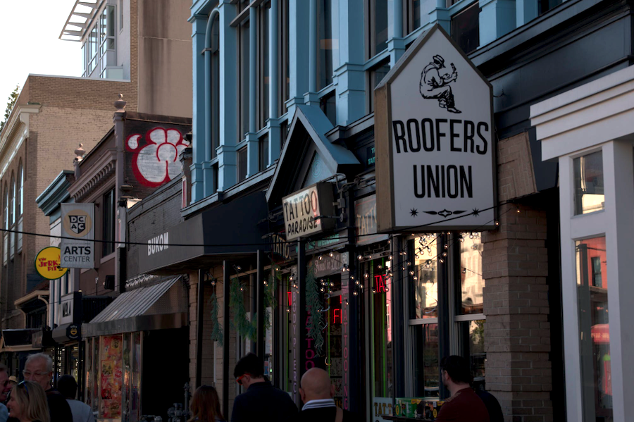 Storefronts and signage, including Roofers Union and DC Arts Center, line a bustling street in Adams Morgan.