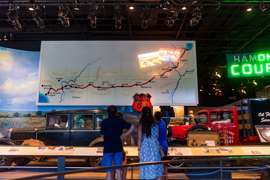 Visitors view a large Route 66 map display with vintage cars and neon signs.