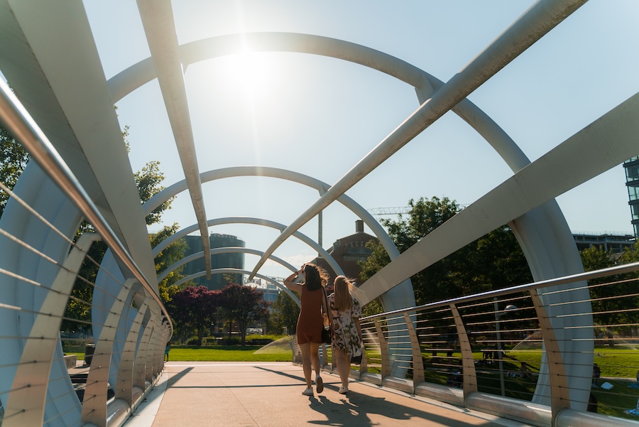 Two people walk across the arched pedestrian bridge at Yards Park as the sun shines brightly overhead.