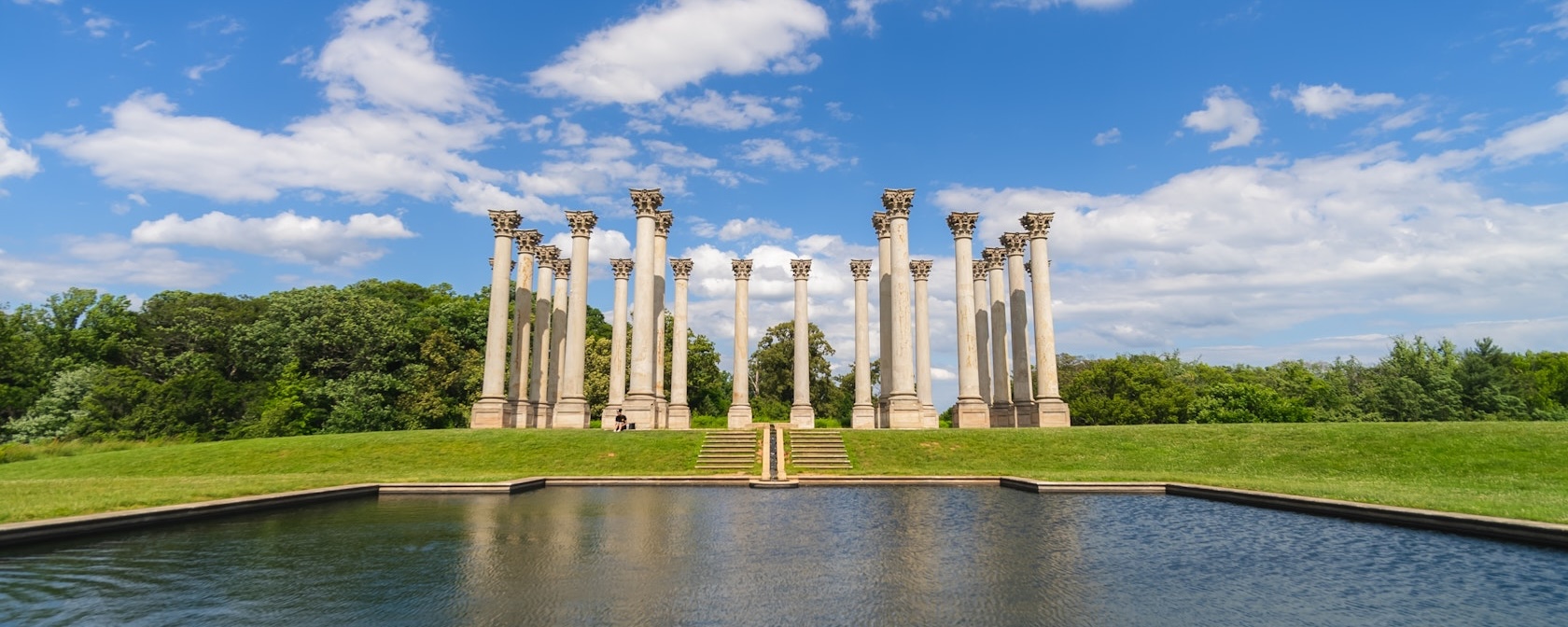 The National Capitol Columns stand tall on a grassy hill reflected in a pond at the U.S. National Arboretum under a bright blue sky.