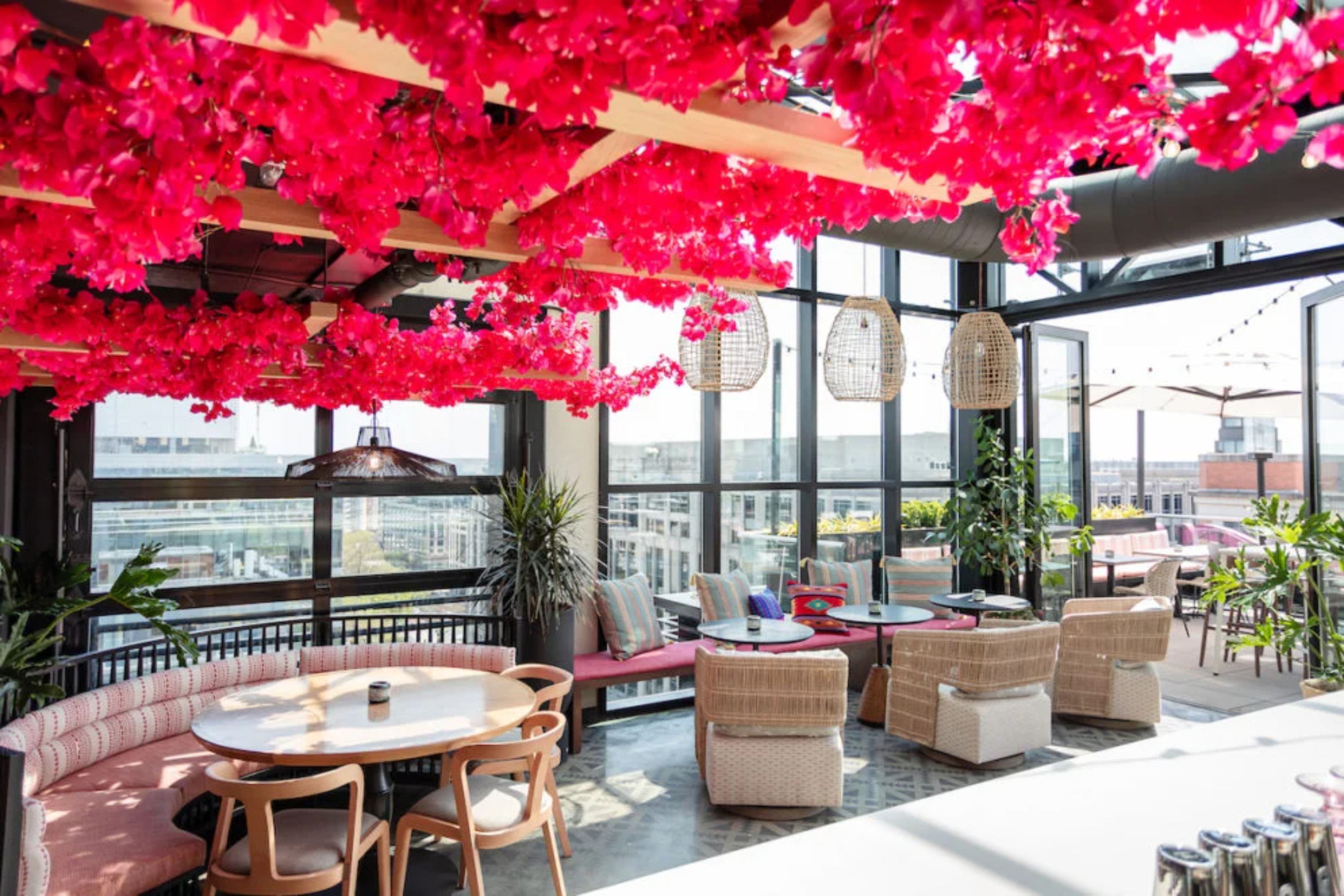 **A bright rooftop lounge filled with natural light features vibrant pink flowers hanging from the ceiling, wicker furniture, lush plants, and cozy seating areas overlooking the city.**