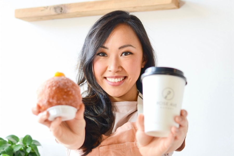 A smiling woman holds up a fluffy, filled donut in one hand and a coffee cup in the other.