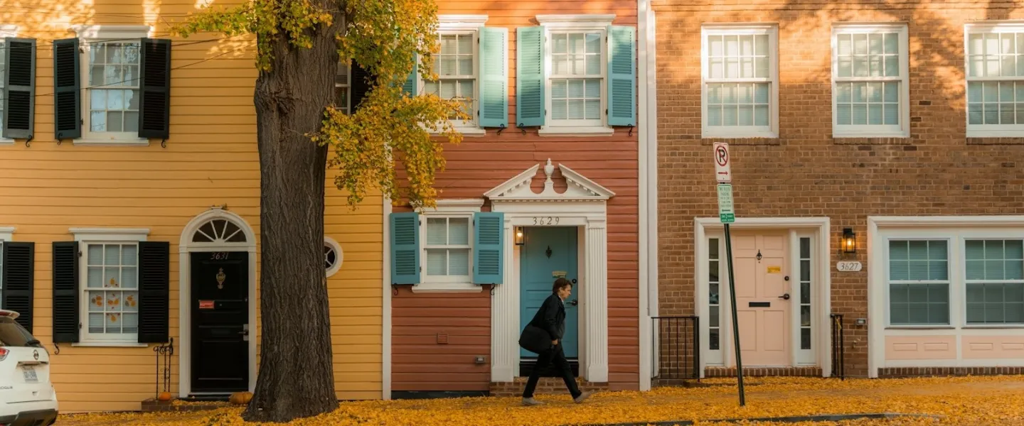 A person walks past colorful historic row houses in Georgetown, their shutters and doors framed by golden autumn leaves.