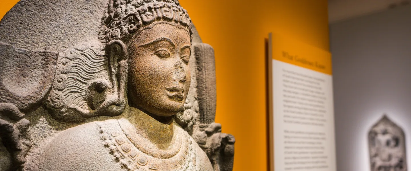 A carved stone Hindu figure is displayed in a museum gallery with warm yellow lighting in Washington, DC.