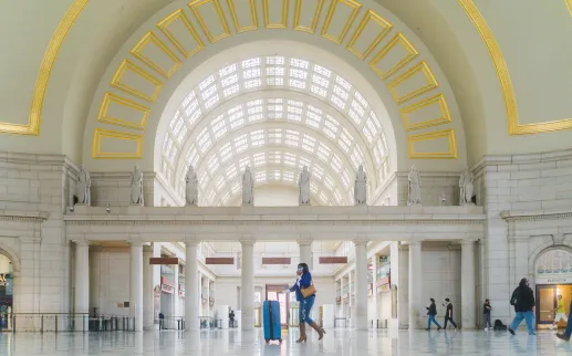  A woman in a blue coat and brown boots walks with a blue suitcase through the grand, arched interior of a spacious train station adorned with statues and large windows.
