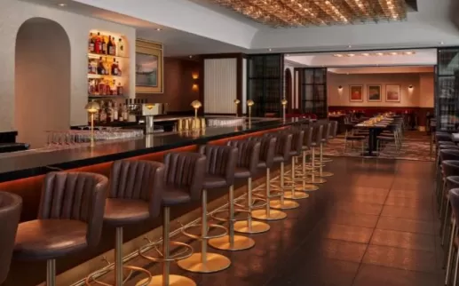 A stylish restaurant and bar with a long, sleek counter lined with leather barstools, illuminated liquor shelves, and a warmly lit dining area with modern decor in the background.