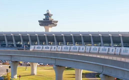 Silver Line Metro train traveling on elevated tracks with Washington Dulles International Airport terminal and air traffic control tower in the background.
