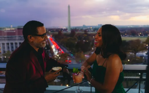 A couple enjoys cocktails on a rooftop terrace with the Washington Monument visible in the background at dusk.