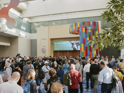 A large crowd of conference attendees gathers in a bright, spacious convention center lobby with colorful signage and modern architectural elements.