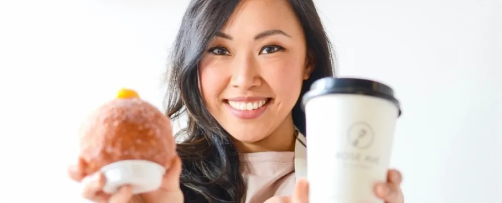 A smiling woman holds up a fluffy, filled donut in one hand and a coffee cup in the other.